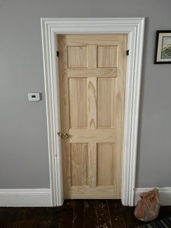 Unfinished wooden door with panel design, framed in white trim against a gray wall.