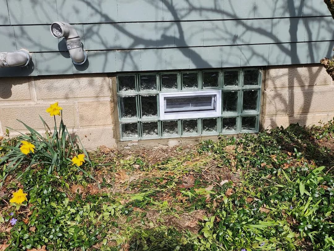 Glass block window with white frame beside yellow daffodils and green foliage.