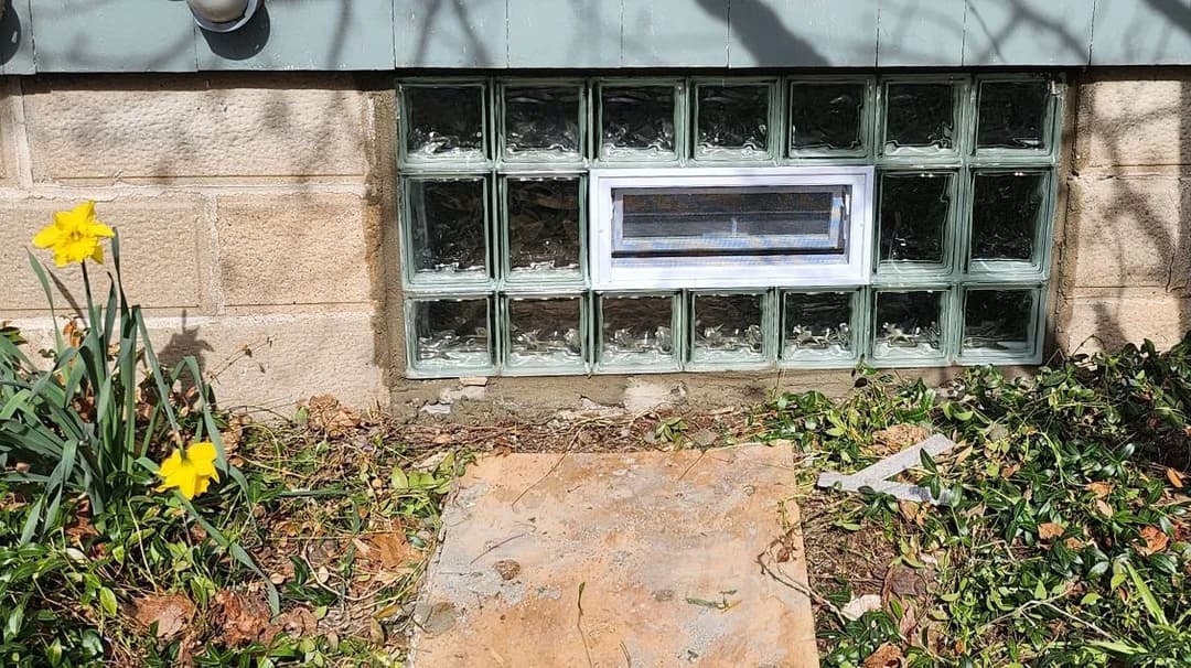 Glass block window in a stone foundation with yellow daffodils and scattered leaves.